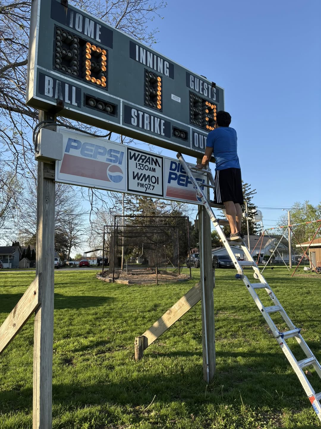 A volunteer on a ladder repairing the scoreboard, with Pepsi and local radio sponsor ads visible
